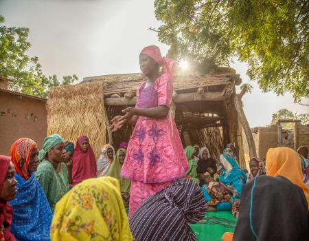 A woman standing in the middle of a group of women gathered sitting on the ground in a settlement