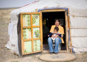 A woman sits in the doorway of a yurt, holding a small child on her lap