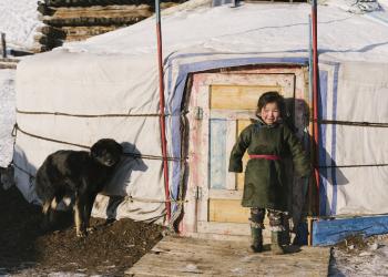 A child dressed warmly in traditional clothing smiles while standing outside a yurt